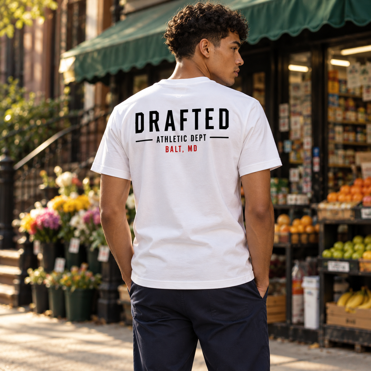 White Drafted Brand Tee — model outside a NYC bodega, back view